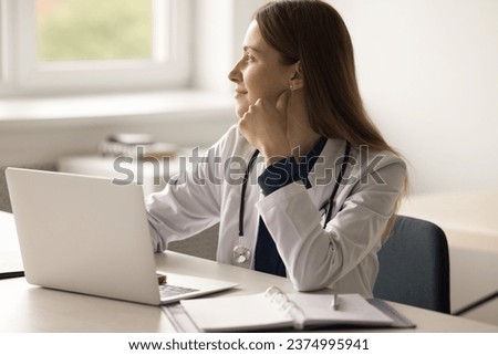 Pensive young general practitioner sits at workplace desk with laptop in hospital, staring out window distracted from work, plan patient treatment, looks dreamy thinking of future and career. Medicine Royalty-Free Stock Photo #2374995941
