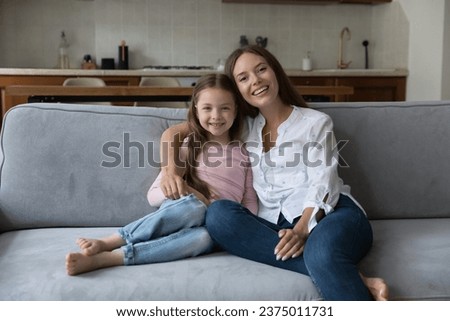 Happy pretty young mom and little daughter kid resting on home couch, hugging, sitting close together, looking at camera, smiling. Mother and kid family portrait. Parenthood, motherhood concept