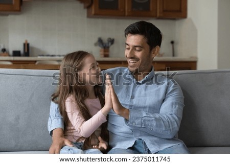 Proud kid girl and happy father giving high five on home couch, hugging, talking, laughing, enjoying leisure. Father giving praise, support to little daughter. Family, parenthood, fatherhood concept Royalty-Free Stock Photo #2375011745