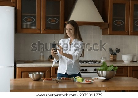 Happy pretty cook blogger girl taking video, picture on mobile phone for social media stories, cooking salad from fresh vegetables, holding smartphone over kitchen table with organic food