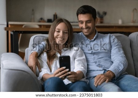 Cheerful happy young couple in love using mobile phone, resting on sofa together, talking on video call, taking selfie picture on smartphone, watching live online media content, enjoying home leisure