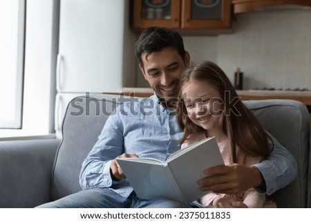 Happy joyful dad teaching cute little daughter to read, hugging kid, showing opening book, resting on couch with child telling fairy tale. Sweet child enjoying studying activity with daddy