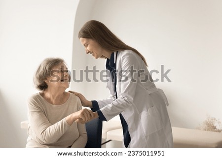 Smiling retired woman visiting doctor in hospital, get good news after medical check-up. Young female general practitioner holds patient hand, touch shoulder, encourages, give psychological support