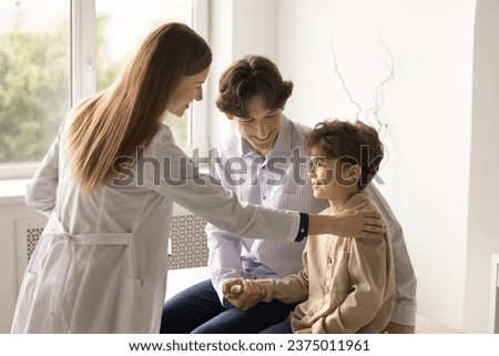 Young female pediatrician in white coat support little boy patient touch kid shoulder, give advice, provides medical assistance. Happy small child enjoy checkup meeting with dad. Check-up, healthcare Royalty-Free Stock Photo #2375011961