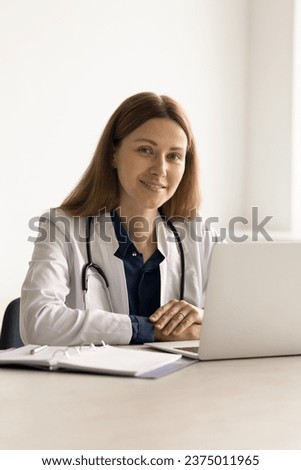 Female therapist in white coat sit at desk in hospital use laptop for work, look at camera, pose at workplace at clinic, advertising medical services. Medicine, professional occupation person portrait Royalty-Free Stock Photo #2375011965