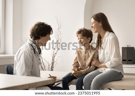 Little cute boy visit pediatrician doctor with loving mom. Friendly male paediatrician in coat hold clipboard writes symptoms, listen to kid patient, provide consultation. Healthcare, medical check-up Royalty-Free Stock Photo #2375011995