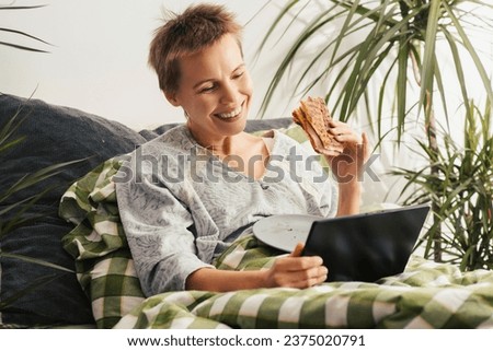 Throughout the day, a woman of middle age, her hair cut short, lies in bed, engrossed in her tablet, eating a sandwich. Apathy, indifference. Bed rotting Royalty-Free Stock Photo #2375020791