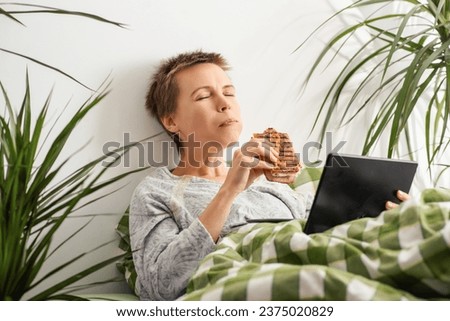 Throughout the day, a middle-aged woman with short hair lounges in bed, glued to her tablet screen and munching on a sandwich. Indolence, passivity. Bed rotting Royalty-Free Stock Photo #2375020829