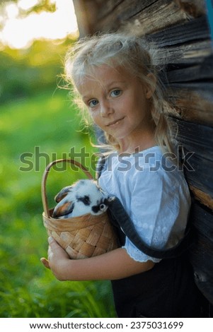 A little girl holds a basket with a bunny, hugs her real pet bunny and learns how to take care of the animal. Child on the street near the house.