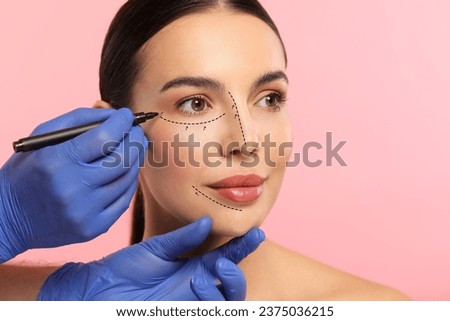 Woman preparing for cosmetic surgery, pink background. Doctor drawing markings on her face, closeup Royalty-Free Stock Photo #2375036215