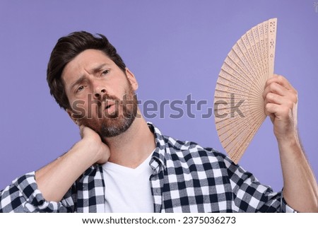 Unhappy man with hand fan suffering from heat on purple background