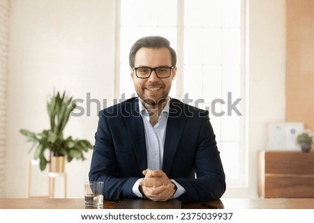 Confident positive handsome businessman in eyeglasses looking at camera, smiling, speaking, sitting at workplace table. Freelance professional man talking on video call. Head shot portrait Royalty-Free Stock Photo #2375039937