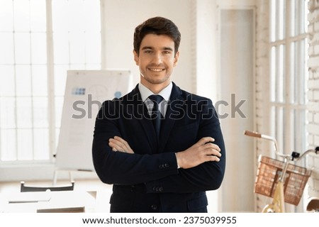 Happy handsome young business leader, entrepreneur man working in millennial office boardroom, standing with arms folded, looking at camera, smiling, enjoying confidence. Professional portrait