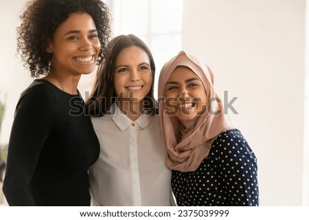 Happy young Muslim professional woman standing close with female office colleagues, smiling, looking away for shooting, hugging wit coworkers, enjoying multicultural corporate friendship