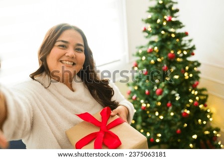 Plus size hispanic woman smiling taking a selfie with her presents next to christmas tree celebrating the holidays looking happy