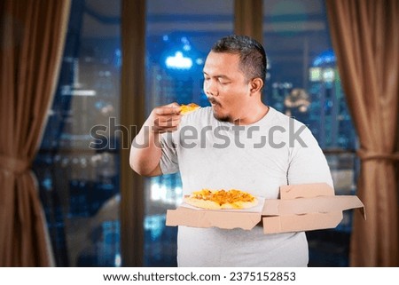 Overweight Asian guy holding a box of pizza while eating it in an high rise apartment building Royalty-Free Stock Photo #2375152853