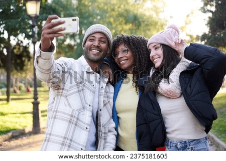 Multiracial group of three friends gathering outdoors, taking a selfie together to post on social media, using a phone. 
