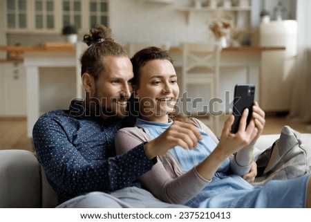 Happy sweet young couple taking selfie picture on mobile phone, watching live media content, talking on video call, using online app on digital gadget, relaxing on couch, enjoying leisure at home