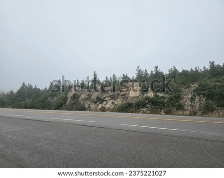 An open road on a foggy day running alongside a rocky cliff.