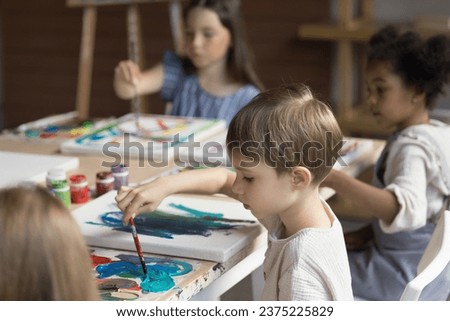 Adorable multiracial children sit at table drawing with paints on canvas. Creative hobby, favourite activity, painting class to nurture kids creativity and imagination, to improve fine motor skills Royalty-Free Stock Photo #2375225829