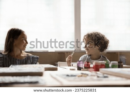 Little pretty girl talking to cute preschooler boy, engaged together in art-class sit at table painting pictures with paints on canvas, enjoy creative activity. Friendship, hobby, talent development