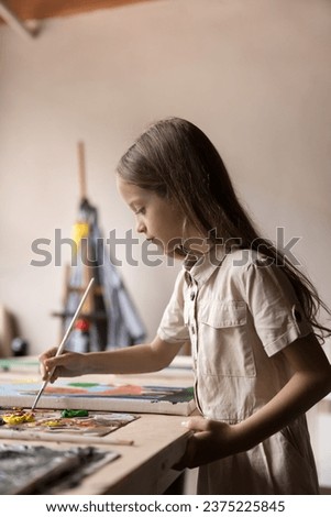 Little girl in casual clothes painting pictures in workshop. Serious kid hold paintbrush drawing with colorful paints on canvas looking focused, enjoy creative hobby standing at table in art studio