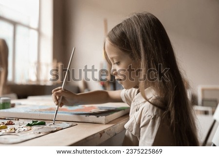 Cute serious little girl painting pictures sit at table in workshop, child holding paintbrush drawing with paints on canvas looking concentrated, enjoy creative hobby in studio. Development, vocation