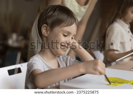 Cute little boy sit at table holding paintbrush makes yellow paint strokes on canvas, smile, enjoy painting group class, close up. Kids development, hobby to improve creative ability, self-expression
