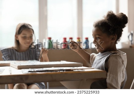 Adorable little African girl holding paintbrush drawing pictures on canvas, sit at table with groupmate, attend art classes or daycare, engaged in favourite creative hobby. Talent development of kids