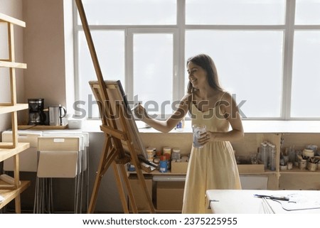 Cheerful attractive female painter holding paintbrush painting on canvas alone in creative art-studio, enjoy hobby or professional occupation, create pictures or personal exhibition smile feel happy