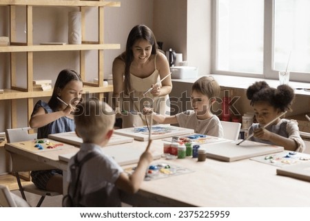 Young artist female teacher lead art-class for little children, multi-ethnic kids drawing pictures with paints on canvas seated at table in art-studio. Talent development, creative hobby, education