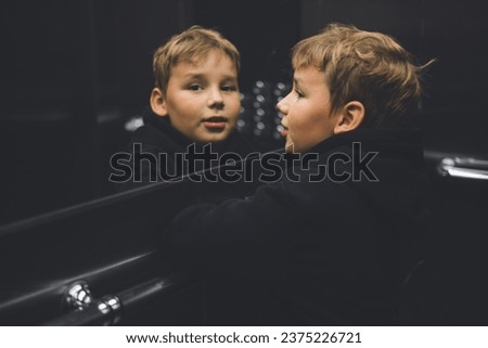 A child goes up in an elevator and looks in the mirror, smiling. A child looks through the elevator mirror into the camera lens with a smile. A handsome boy looks into a mirror in an elevator.