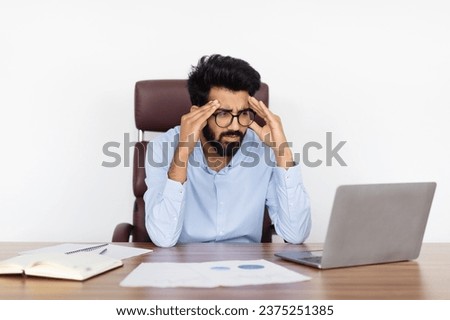 Stressed millennial eastern businessman working at office, sitting at table over white wall background, looking at laptop computer screen and touching his head, have problems with business, crisis