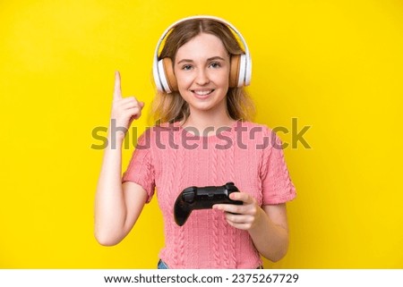 Blonde English young girl playing with a video game controller isolated on yellow background pointing up a great idea