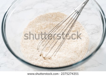 Mixing dry ingredients with a hand whisk in a large glass mixing bowl to bake mini vanilla cupcakes with ombre pink buttercream frosting. Royalty-Free Stock Photo #2375274351
