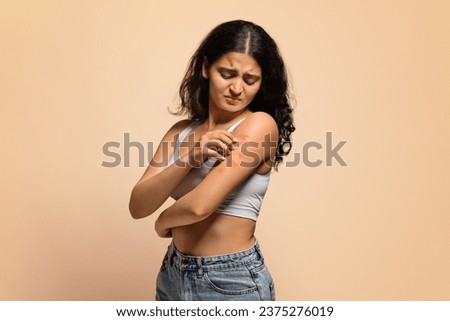 Dermatitis, eczema, allergy, psoriasis concept. Annoyed young indian woman scratching irritated skin on arm, eastern female having itching rash on body, standing on beige studio background Royalty-Free Stock Photo #2375276019