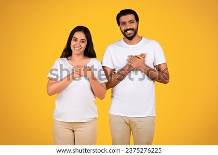Loving couple placing hands on chest, over heart, isolated on yellow studio background. Faces exhibit trust, commitment, and shared happiness in a heartfelt moment, gratitude, thanks Royalty-Free Stock Photo #2375276225