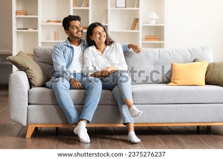 Joyful Indian couple, comfortably sitting on a sofa in their homey space, sharing a light, dreamy moment while gazing aside and sharing tender smiles, free space