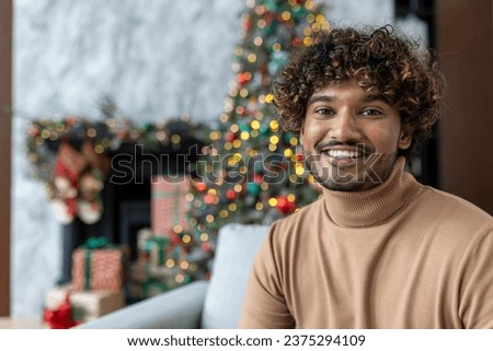 Christmas portrait of young happy man, Hispanic man smiling and looking at camera, happy and celebrating Christmas sitting on sofa in living room near decorated Christmas tree.