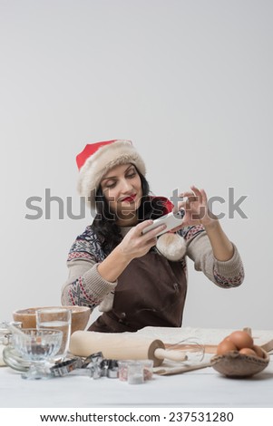 Happy young housewife making christmas cookies in kitchen and taking pictures on smartphone