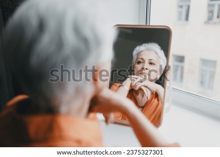 Senior elderly female with gray hair and wrinkles looking in mirror, upset with aging process of her body, reflecting upon her youth and young facial skin, missing her past beautiful appearance Royalty-Free Stock Photo #2375327301