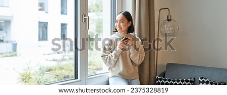 Beautiful young asian woman sitting near window and drinking her coffee, holding espresso cup and looking outside with relaxed, smiling face expression. Royalty-Free Stock Photo #2375328819
