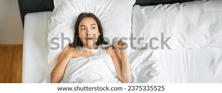 Portrait of asian woman lying in bed with shocked face, looking startled and upset, gasping from smth, staying alone in her bedroom. Royalty-Free Stock Photo #2375335525