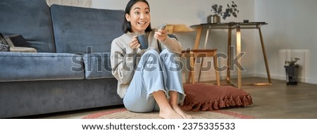 Portrait of girl watching television at home, sits on floor near sofa, holds remote and changes channels.