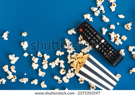 Remote control and black and white stripped box full of popcorn on a blue background. Watching TV at home. Entertainment concept. Tv shows or movie night background with copy space.