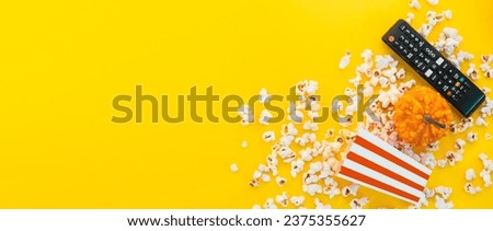 Remote control and red and white stripped box full of popcorn on a yellow background banner. Watching TV at home. Entertainment concept. Tv shows or movie night background with copy space.