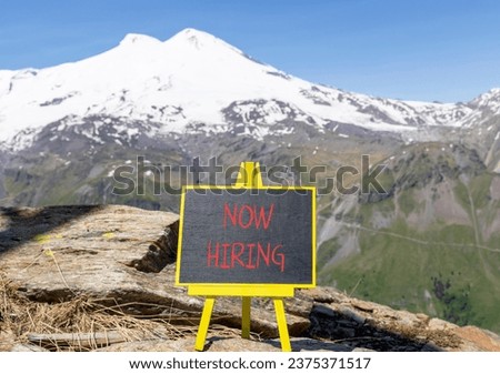 Now hiring symbol. Concept words Now hiring on beautiful black chalk blackboard. Beautiful mountain Elbrus blue sky background. Business marketing, motivational now hiring concept. Copy space.