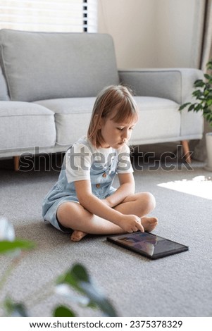 A cute preschool girl looks at the tablet screen while sitting on the floor in the room. Children's educational apps for phone and tablet. Children and digital technologies.