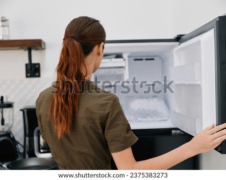 A woman in the kitchen of her home opened an empty freezer with ice inside, home refrigerator, defrosted, view from the back. Royalty-Free Stock Photo #2375383273