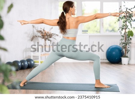 Proficient young female yoga instructor doing warrior pose variation in light sports room